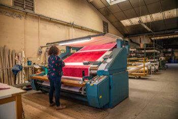 Une visiteuse examine attentivement les mètres de tissu, déroulés avec soin, lors d'une visite guidée dans la manufacture. Elle observe la matière et les motifs avec curiosité et précision, illustrant l’authenticité et le savoir-faire textile de Beauvillé.