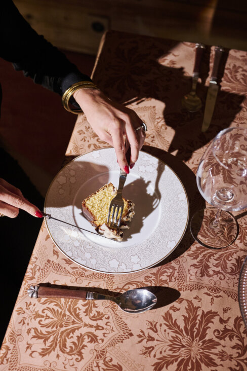 Vue de haut d’une table dressée avec la nappe Topkapi moka. On y voit des mains coupant une part de gâteau à la vanille avec un couteau et une fourchette, dans une ambiance chaleureuse et élégante.