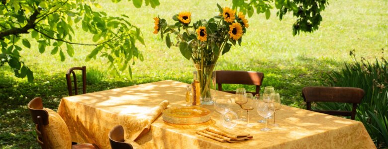 Nappe Topkapi coloris miel, dressée au milieu du jardin sous les arbres. Un bouquet de tournesols orne la nappe rappelant la chaleur et la luminosité du coloris miel.